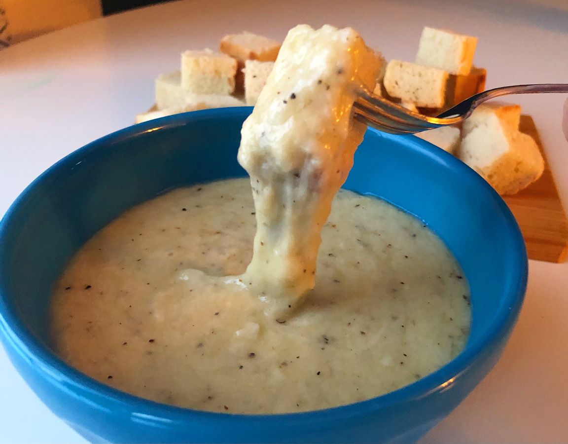 Cheese fondu from The Bread Company in Urbana is in a blue bowl. A bread cube has been dipped by a metal fork in the cheese, and a big thick strand of melted cheese connects the bread to the fondu. Photo by Alyssa Buckley.