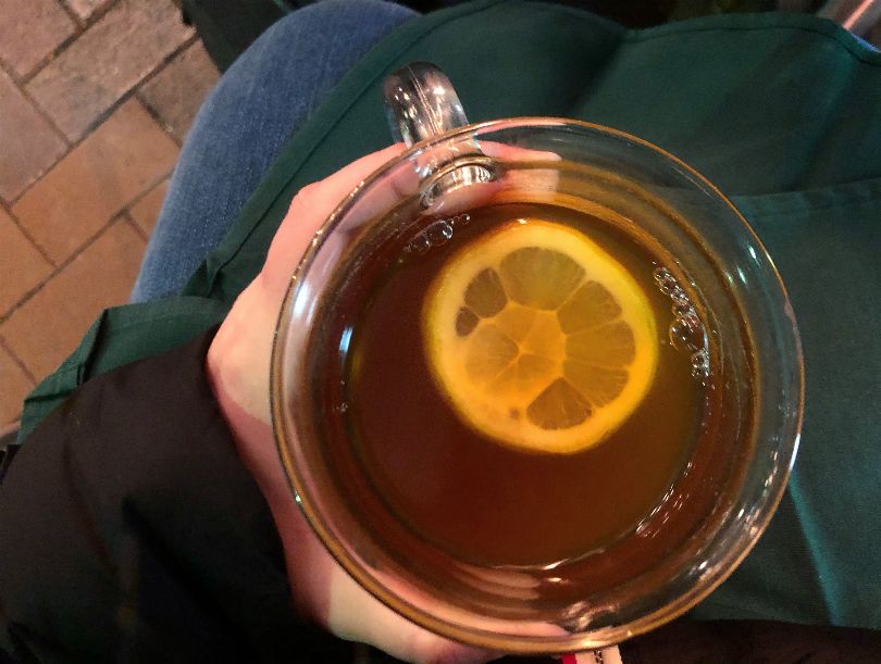 An overhead shot of the author's hot toddy. The lemon circle is floating in a brown drink in a clear glass mug above the author's jeaned lap. Photo by Alyssa Buckley.