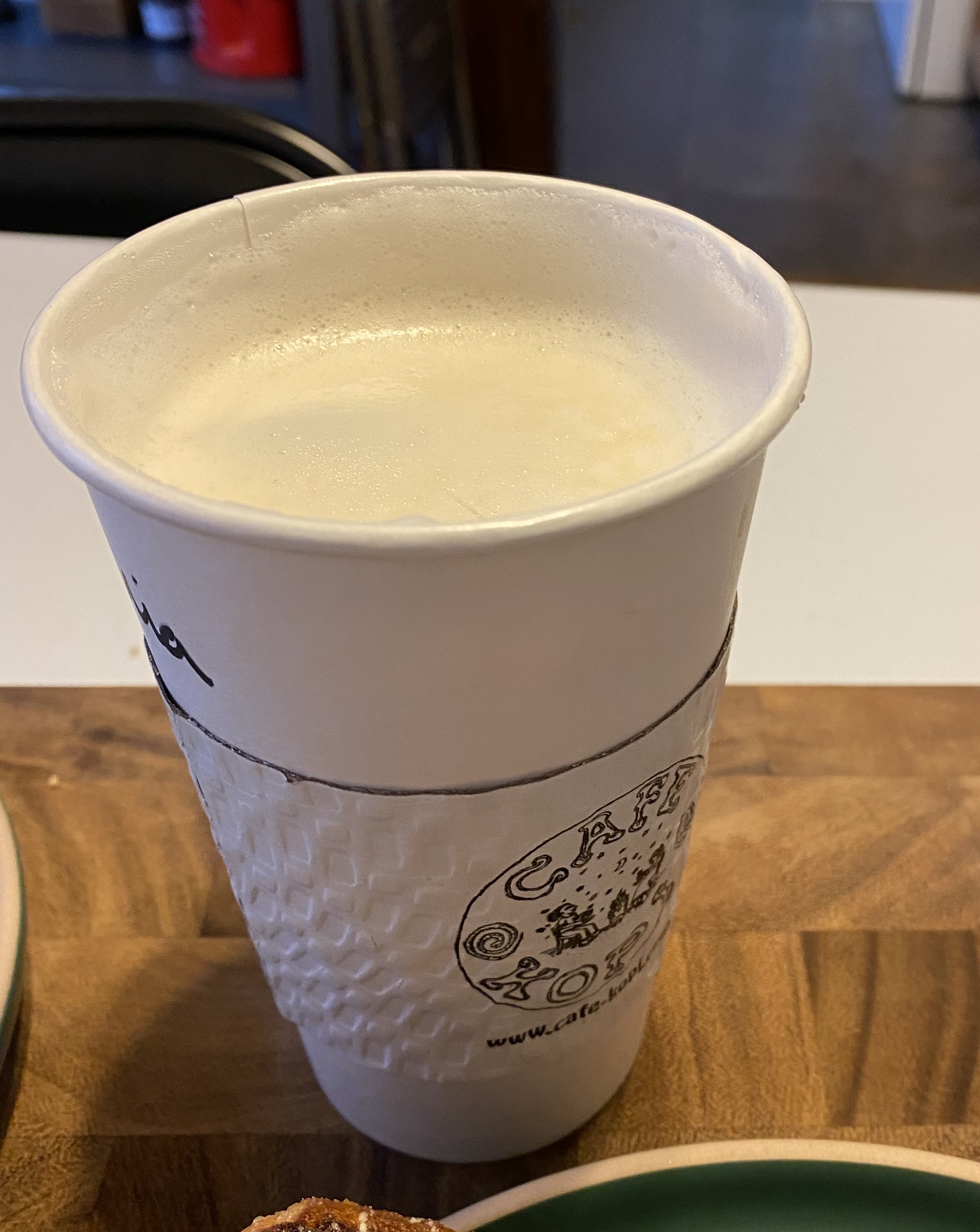 A paper coffee cup with a paper sleeve with Cafe Kopi's name and logo on it hold a hot latte with a white foamy top. Photo by Mia Hanneken.