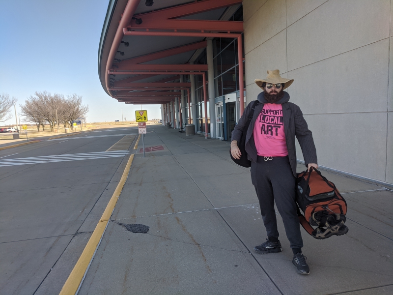 The writer is standing on the sidewalk near the airport entrance facing the camera. He is wearing a floppy hat and sunglasses and holding his bags. Photo by Andrea Black.