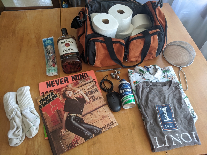 An assortment of items are on a light wood table: An orange duffel bag with several rolls of toilet paper, socks, a bottle of bourbon, a model airplane, a black computer mouse, a Tanya Tucker album, a can of Barbasol, and two shirts. Photo by Andrea Black.