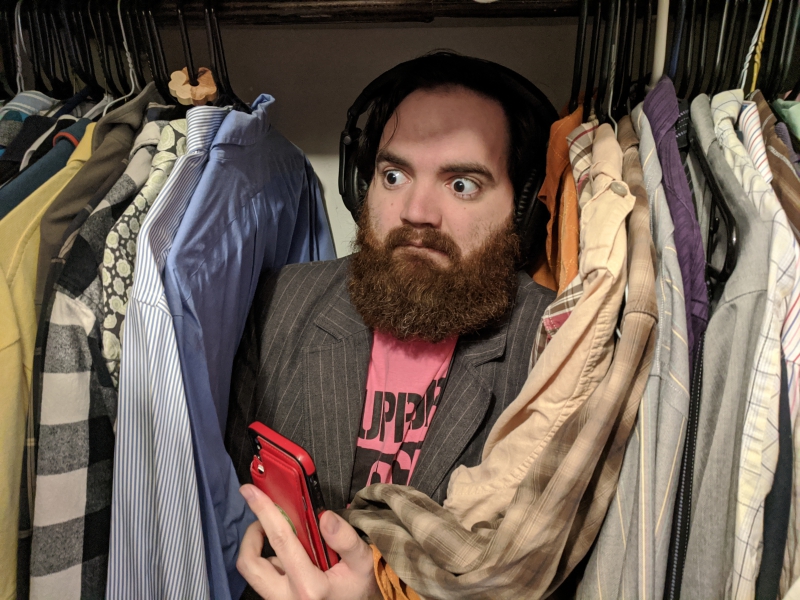 The writer is wedged between shirts hanging in a closet. He has large black headphones on and is holding a phone. Photo by Andrea Black. 