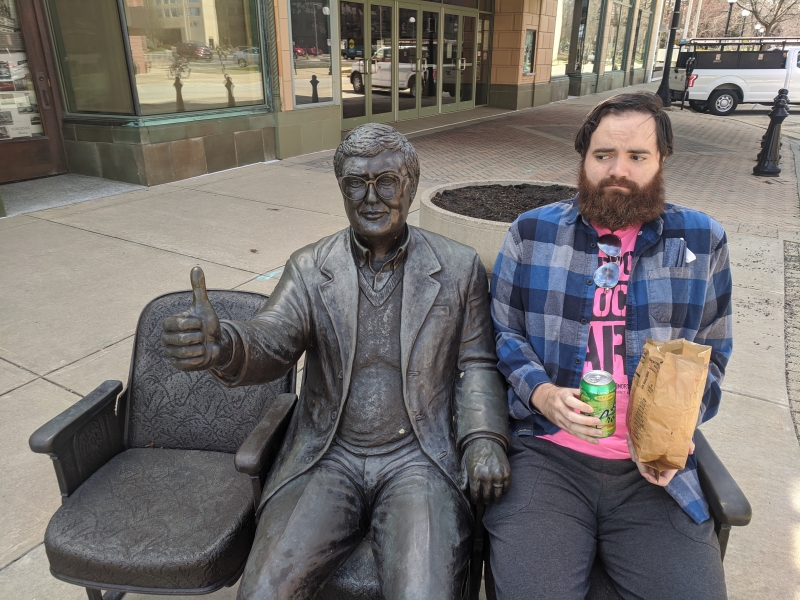The writer is sitting in a seat next to a statue of Roger Ebert. He is holding a La Croix and brown paper bag. Photo by Andrea Black.