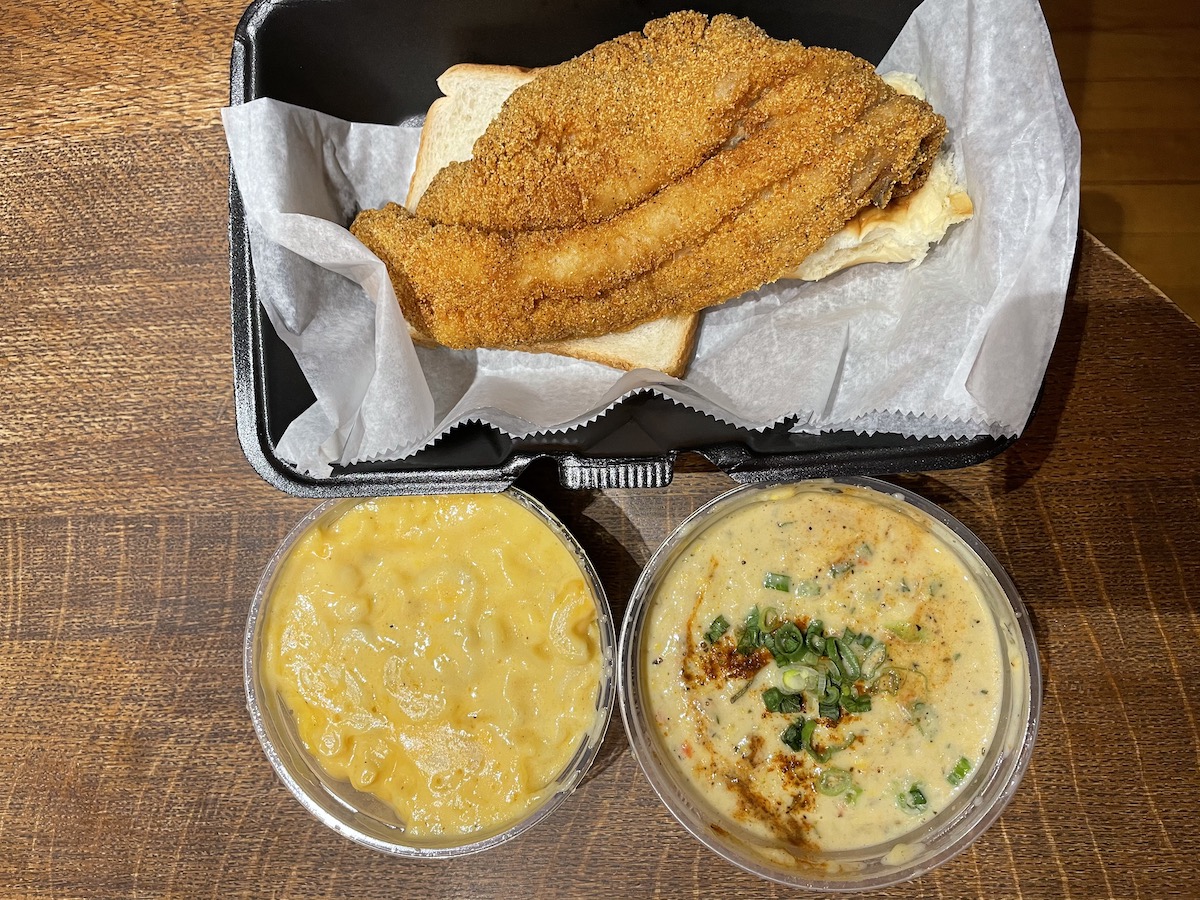 An overhead photo of a catfish dinner from Neil St. Blues with a large fried catfish in parchment paper in a black takeout container. Below is a cup of yellow macaroni and cheese and beside it is a a plastic cup of crab bisque. Photo by Anthony Erlinger.