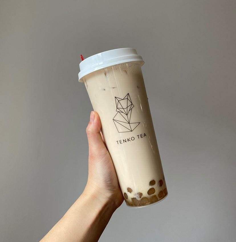 A white hand holds a large boba oolong tea from Tenko Tea in Champaign. The tea is a brownish gray, and the clear cup shows the boba at the bottom of the drink. On the outside of the cup, there is a thin lined fox art with the name of the tea shop. Photo from Tenko Tea Champaign's Facebook page.