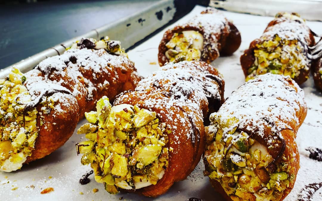 Six fresh cannoli sit on a tray. The ends are coated in chopped pistachios. The cannoli are finished with a drizzle of chocolate and a sprinkle of powdered sugar. Photo by Jordan Baldarotta.