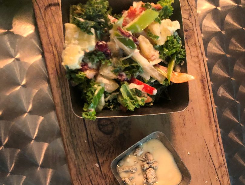 A kale-pineapple slaw sits in a metal square bowl beside a mini cup of a yellow-ish blue cheese sauce on a wooden platter. Photo by Alyssa Buckley.