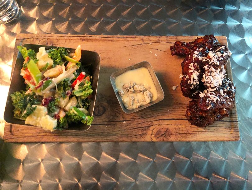 An overhead shot of the wings platter on a long, rectangular piece of wood. On the left, there is a kale-pineapple slaw, in the middle, a miniature dipping sauce, and on the right, six jerk wings covered in sauce on a metal outdoor table. Photo by Alyssa Buckley.