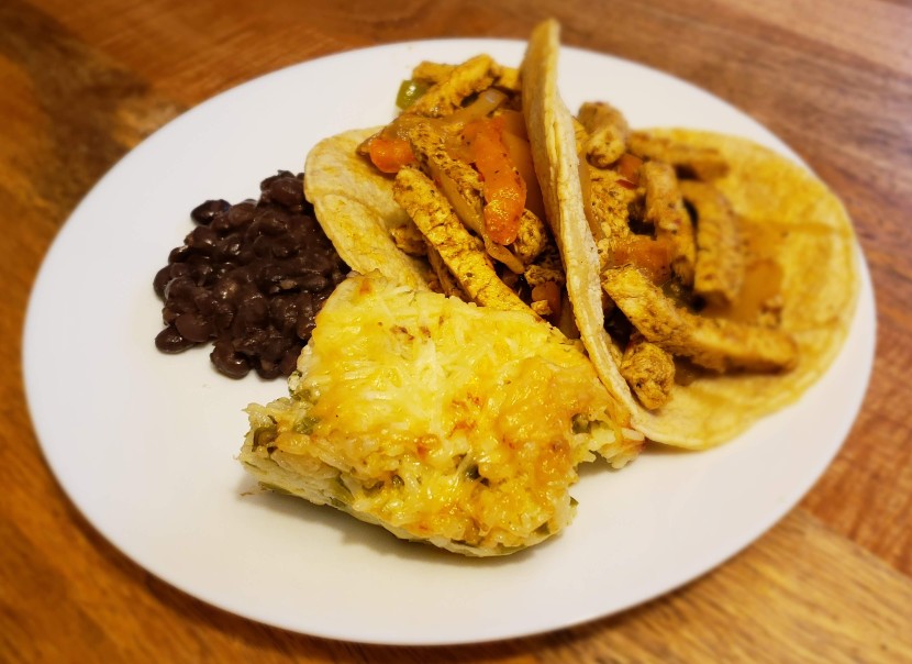 A plate of two vegetarian chicken fajitas made from soy curls and bell peppers served on corn tortillas. Next to the fajitas is a serving of black beans and a square of cheesy, jalapeno and white rice casserole. The round white plate is shown on a wood table background. Photo by Sara Ressing. 