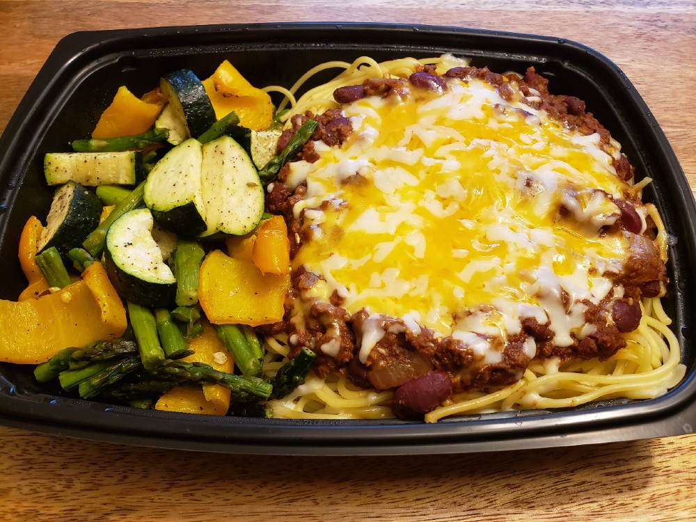 Photo shows two servings of a Piato meal in a black reusable take out container. A light brown table is visible in the upper portion of the picture. On the right side is a mixture of roasted vegetables including half moons of zucchini, green spears of asparagus and sliced orange bell peppers. To the left is a large portion of spaghetti topped with a bright red bean chili and covered in melted cheese. Photo by Sara Ressing. 