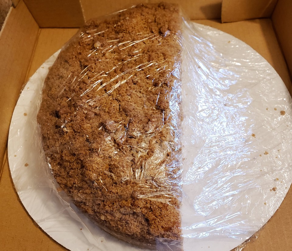  A view of Piato's half-pan of Bailey's coffee cake. THe half-moon cake is shrink wrapped on a white cardboard cake round and delivered in a brown box. The photo shows the top of the coffee cake, which is covered in a brown-sugar crumble topping. Photo by Sara Ressing