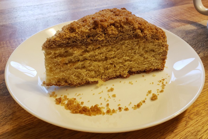 A side view of  a slice of Bailey's coffee cake on a white plate. The coffee cake has a brown topping over a yellow cake base. There are crumbs of cake and topping on the plate. Light is reflected on the plate and brown table background. Photo by Sara Ressing. 