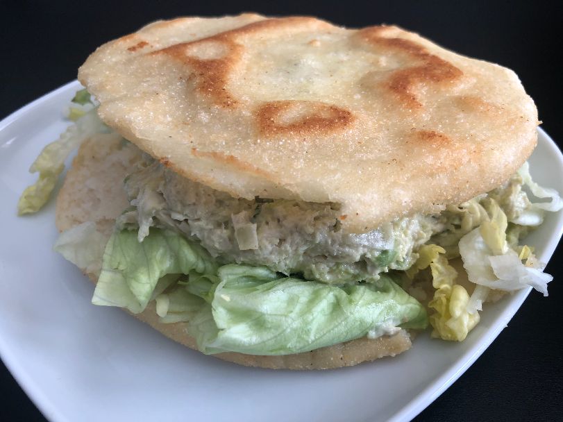An image of arepas from El Paraiso shows grill marks on the thin bun with a generous portion of a creamy chicken filling with another bun below. The arepas sits on a white plate on a black table. Photo by Alyssa Buckley.