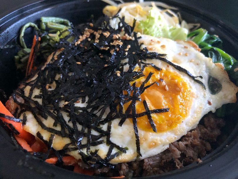 Bibimbap from Masijta Grill in Urbana sits in a black plastic container. The egg on top is perfectly fried with black strands of seaweed overtop. Photo by Alyssa Buckley.