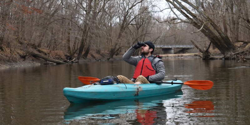 A man in an orange life vest is sitting in a light blue kayak. The kayak is floating on a river, lined with bare branched trees. He is looking through binoculars. Photo provided by Kingfisher Kayaking.