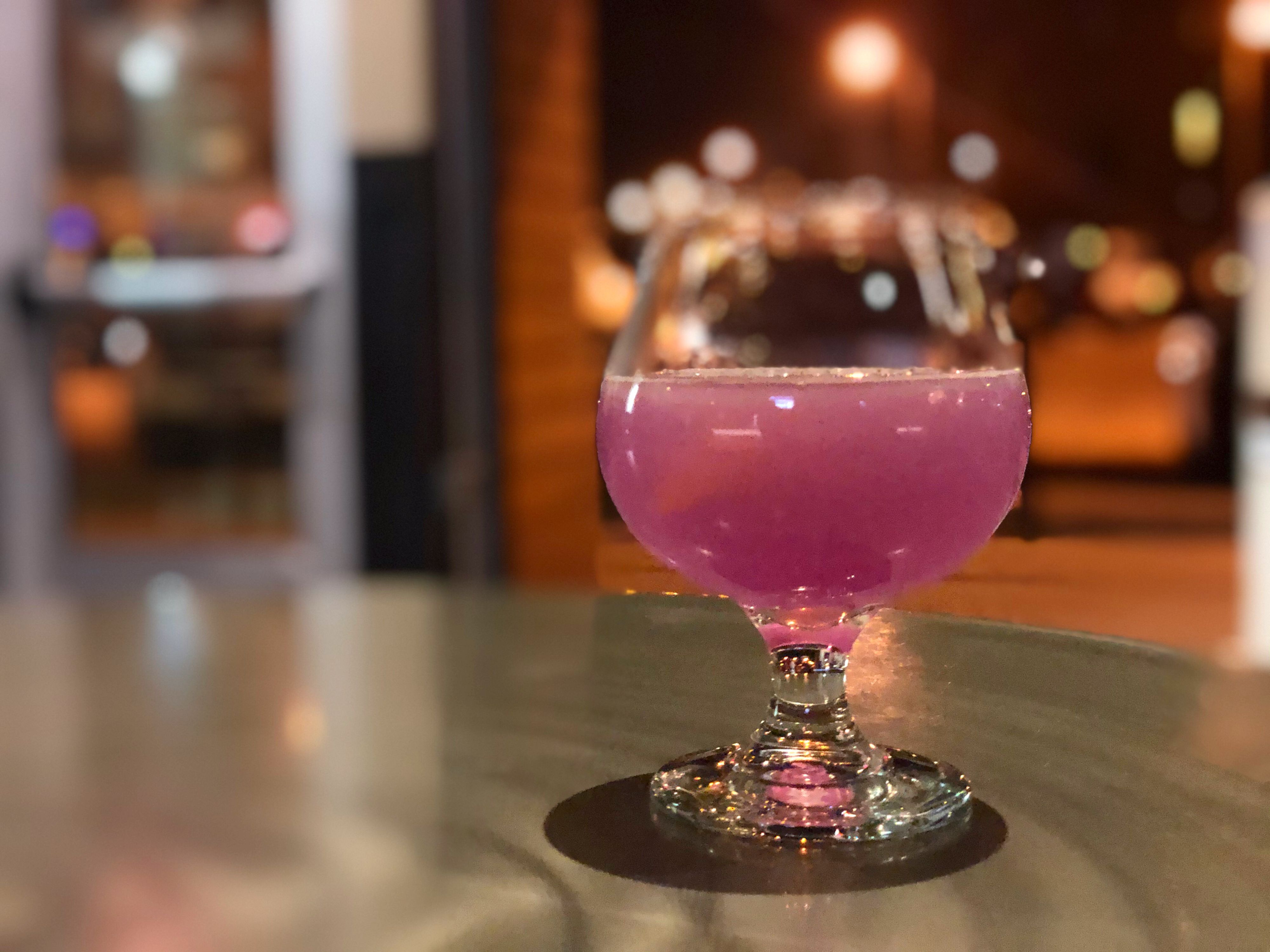 A purple cocktail sits in a snifter glass on a silver table inside Collective Pour. In the blurred background, there are parked cars and the lot beside Neil Street in Downtown Champaign. Photo by Alyssa Buckley.