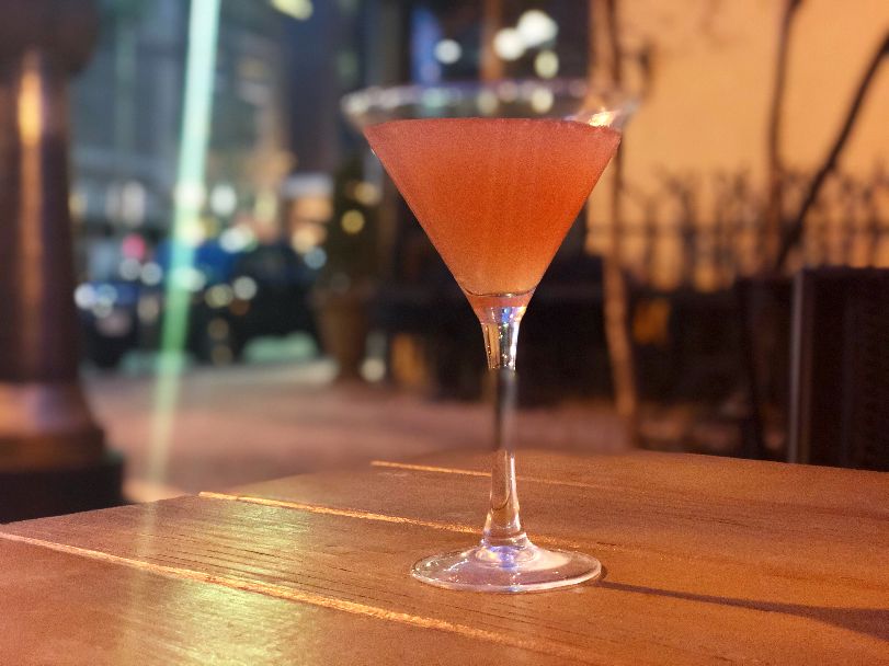 At night, there is a pink cocktail in a martini glass on an outdoor picnic table. Shimmering, blurred street lights and string lights are in the background. Photo by Alyssa Buckley.