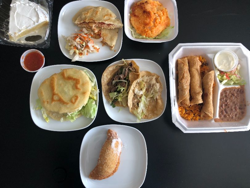An overhead image of the author's order. In the top left, there is a slice of white cake beside pupusas, tacos, llapingachos, flautas, arepas, and a dessert enchilada. Photo by Alyssa Buckley.