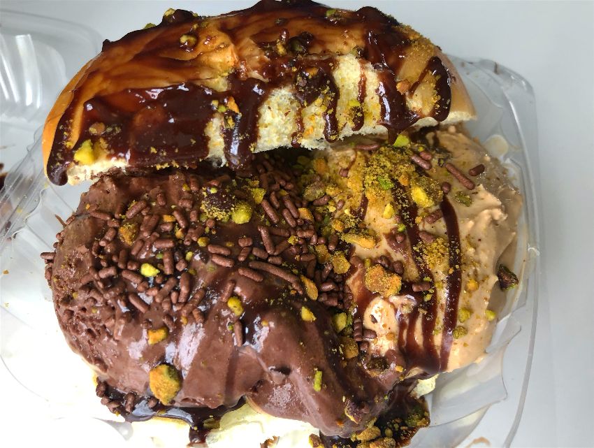 Baldarotta's gelato brioche sits in a plastic clamshell container. Inside a brioche bun are two scoops of gelato, dark chocolate on the left and caramel on the right. There is a lot of chocolate syrup covering the entire dessert with crushed pistachios and chocolate sprinkles in the middle. Photo by Alyssa Buckley.