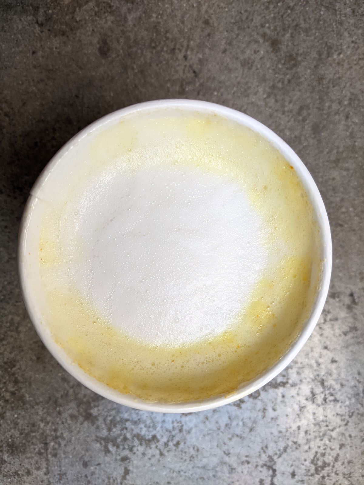 Golden Milk Latte from Common Ground Food Co-op. A top view of a cup of white frothy latte with yellow edges. The cup sits on a grey concrete floor. Photo by Tias Paul.