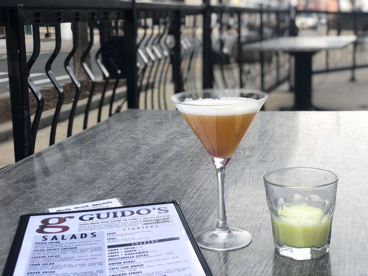 On a silver metal table at Guido's in Downtown Champaign, there are two drinks: a yellow martini and a greenish yellow shot with a laminated menu beside it. Photo by Alyssa Buckley.