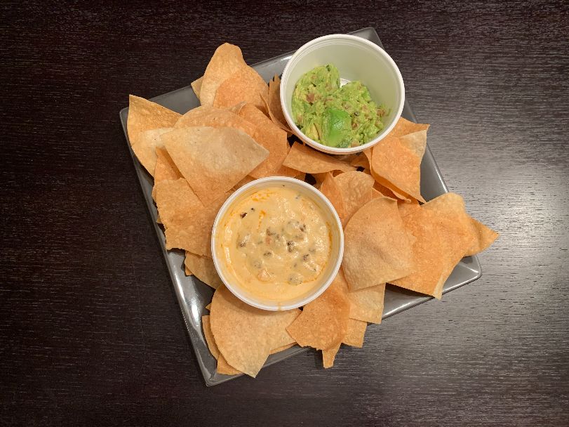 An overhead photo of chips and two dips on a wooden table. The chips are yellow, triangular corn chips with a Styrofoam cup of queso and a cup of guac. Photo by Stephanie Wheatley.