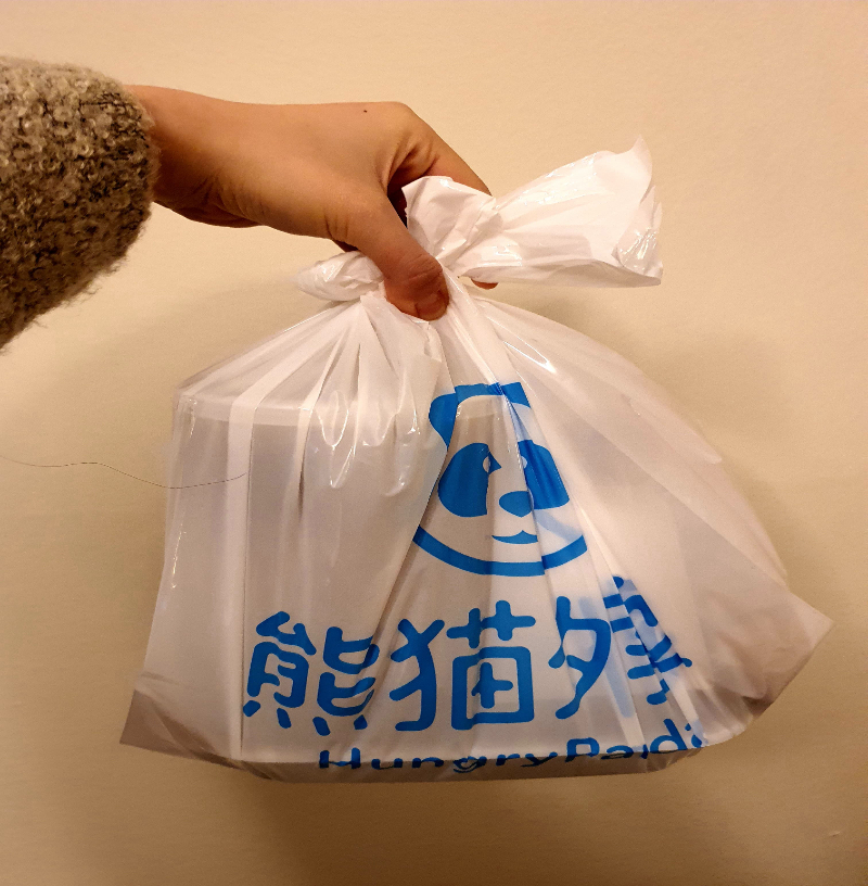 A plastic takeout bag is held by a hand with a brown, fuzzy sweater. The bag has dark blue Chinese writing and a smiling panda. Photo by Da Yeon Eom