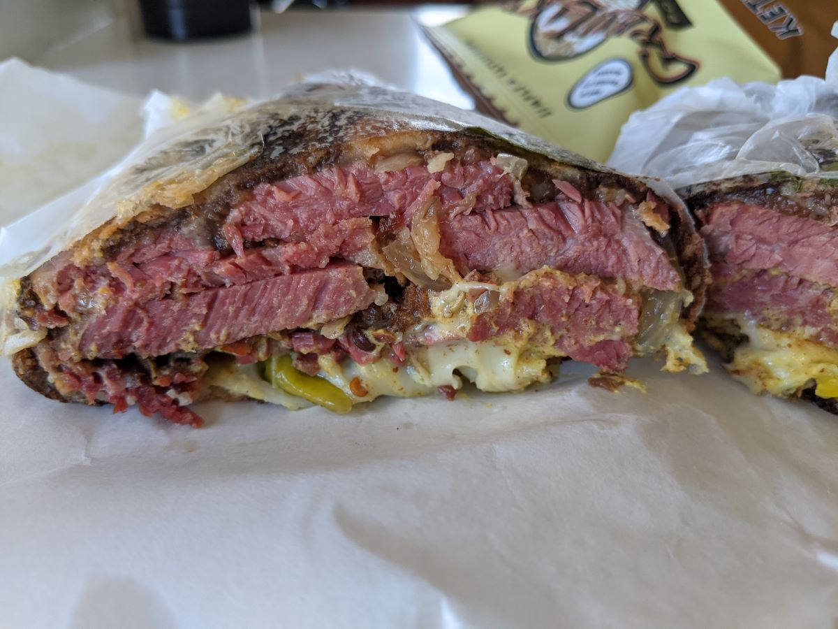 Pastrami and Pepperoncini Melt from Baldarotta's. A cross-section view of a sandwich with thick slices of red/pink pastrami, green pepperoncini, and melted cheese.  In the background (blurry), is a beige bag of chips. Photo by Tias Paul.
