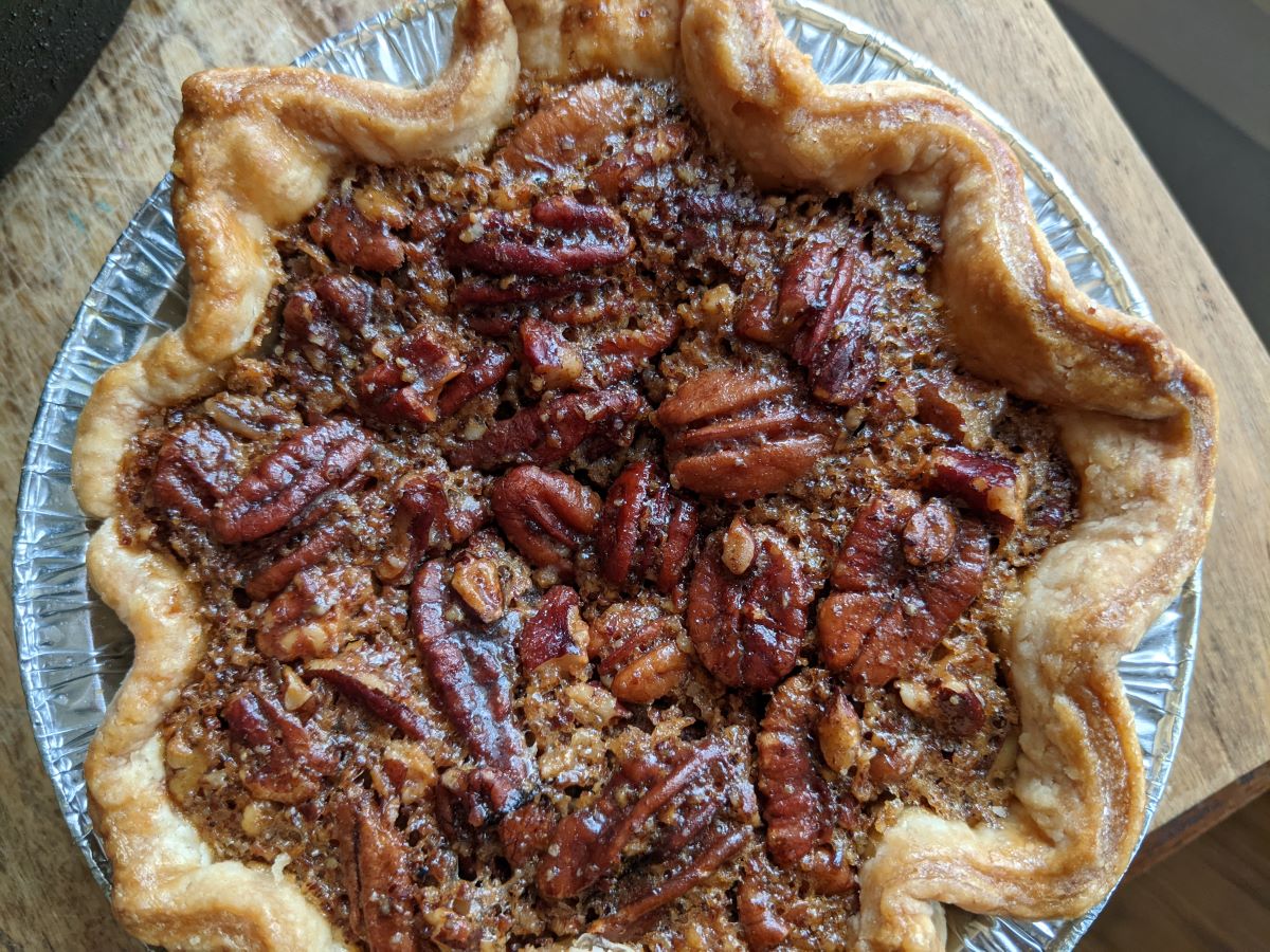 Pecan Pie from Brit's House of Sweets. A round pecan pie with crimped edges set in an aluminum pan. The top (pecans) is shiny mahogany brown. Photo by Tias Paul.