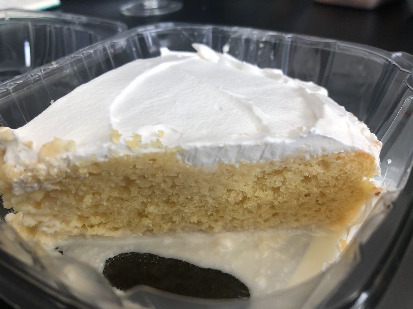 A large slice of tres leches cake sits in a plastic clamshell container with very white frosting and a light yellow cake with dripping leches sauce on the bottom of the container. Photo by Alyssa Buckley.