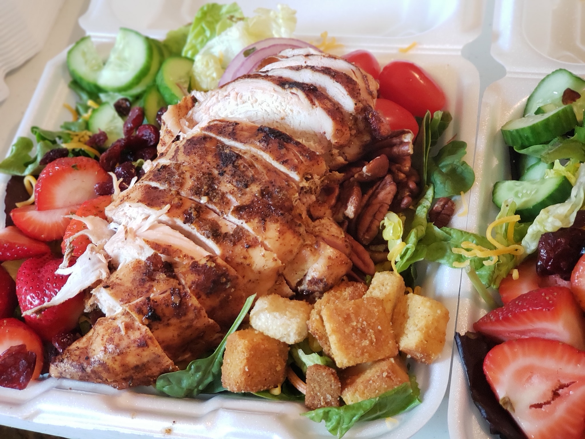 A huge smoked chicken breast is on top of a salad in a styrofoam container with strawberries, almonds, croutons, and cucumbers. Photo from Dis N' Dat BBQ Facebook page.