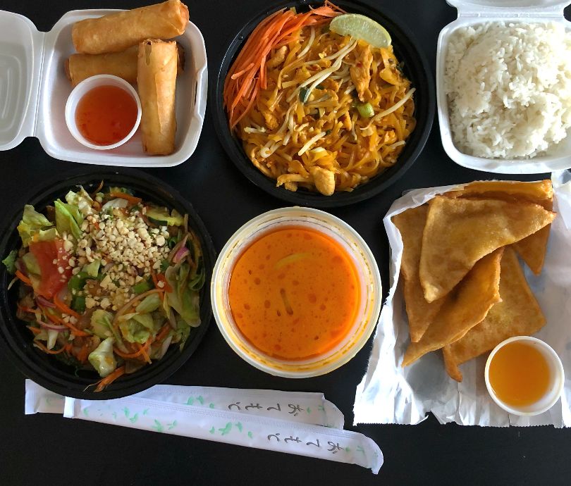 An overhead photo of six dishes from Sticky Rice in Champaign. From the top left, there is a square styrofoam container with three egg rolls and a plum dipping sauce. Then, a circular takeout container of chicken pad Thai with a lime wedge, beside a styrofoam container of steamed white rice. On the bottom left, there is a yum nuau salad, a circular container of curry, and on the right bottom six crab rangoons in a white paper bag. Photo by Alyssa Buckley.