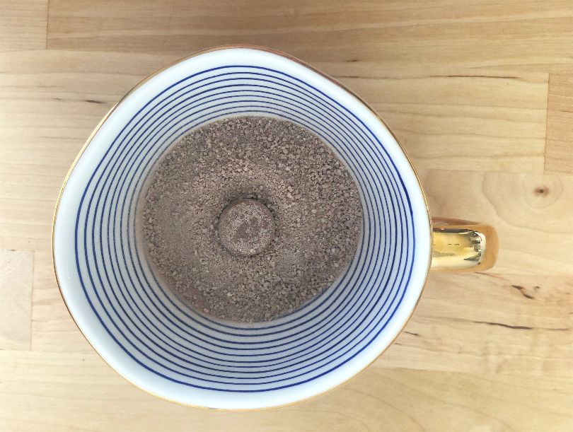 An overhead view of a mug with blue lines on the white interior with a golden handle. The mug has a layer of cocoa powder and a chocolate circle in the center. Photo by Alyssa Buckley.
