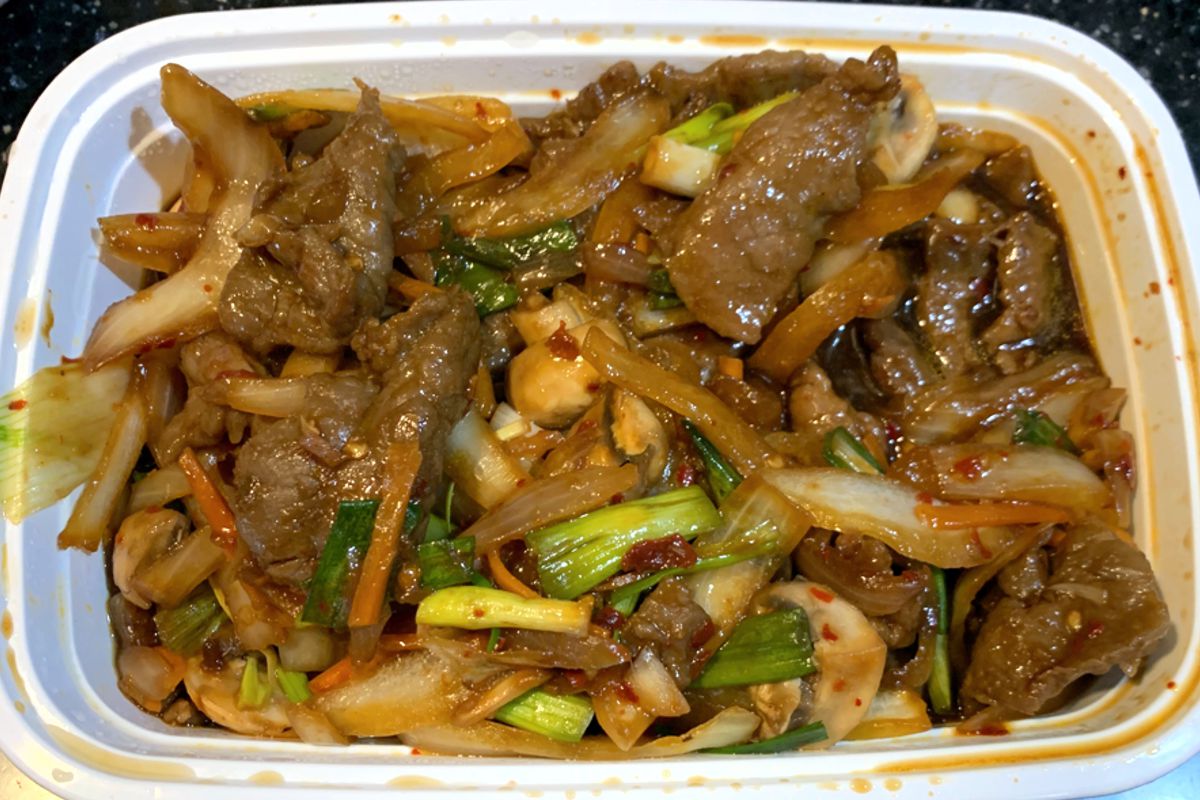 A white to-go container is filled with a mixture of large pieces of brown beef, cooked onions, mushrooms, small slices of carrot, and green onions. The food is covered in a thin brown sauce. Photo by Megan Friend.