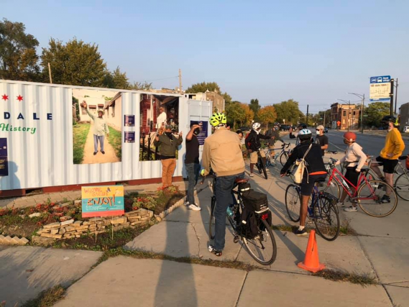 A group of people on bicycle are gathered on a sidewalk. They are looking at a display of photos. Photo from Organic Oneness Facebook page.