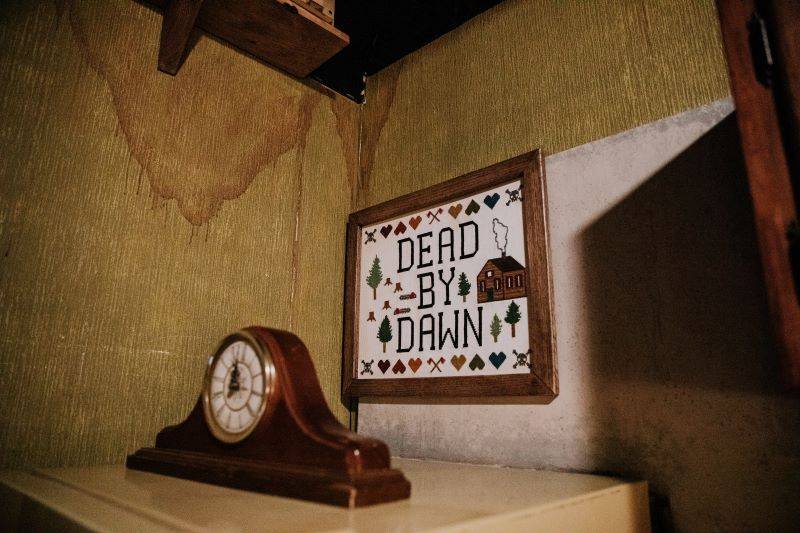 A creepy corner of Revenge of the Cabin, an escape room in Urbana, Illinois, has a shelf with water damage stains, a wooden clock set to midnight, and a framed cross-stictch art reading. Photo by Anna Longworth.