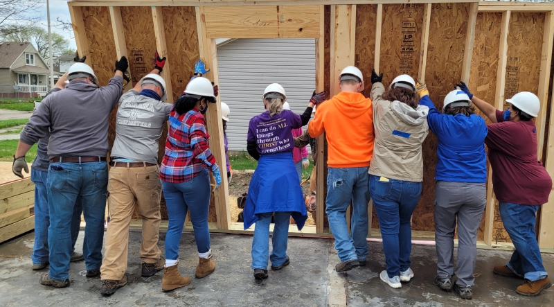 A row of nine people are pushing the wall of a home to standing. They are wearing white hard hats and facing away from the camera. Photo provided by HHCC.