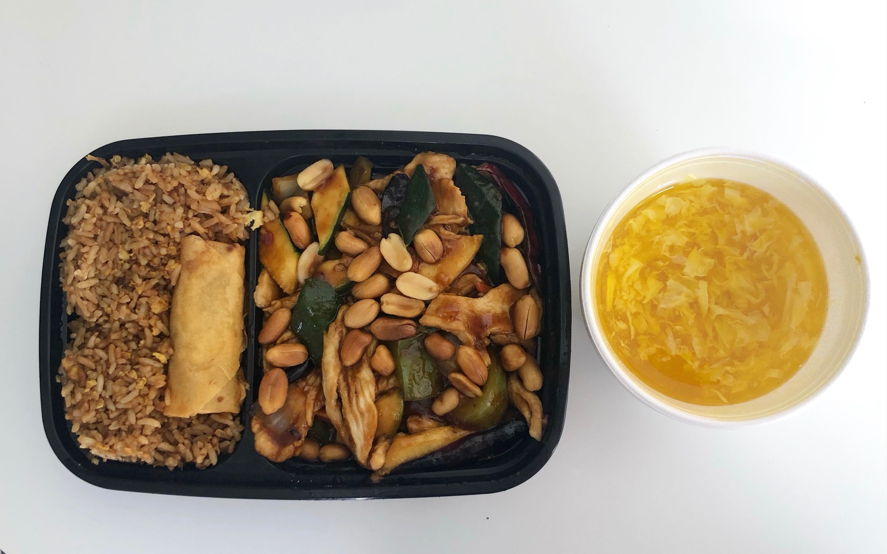 An overhead photo shows a princess chicken lunch dish from Rainbow Garden. In a black takeout container, there is a portion of brown fried rice with a small egg roll on top beside a portion of peanut topped chicken entree. There is a plastic takeout bowl of yellow egg drop soup on the white table. Photo by Alyssa Buckley.