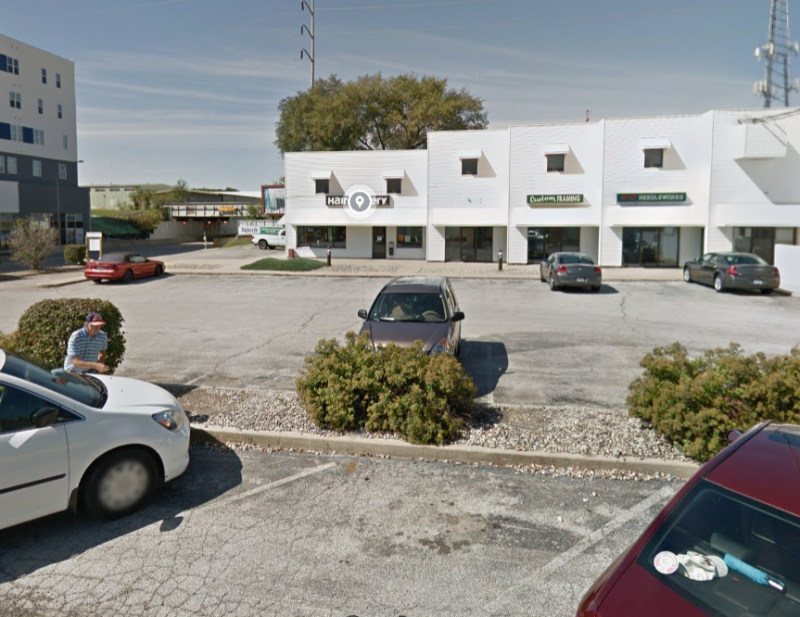 A parking lot with a few cars in parking spaces. On one side there is a building with white siding and multiple businesses. Screenshot from Google Maps.