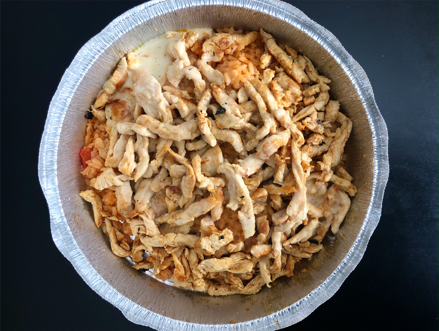 In a metal pie pan, there is a lot of thinly sliced chicken over Mexican rice on a black table. Photo by Alyssa Buckley.