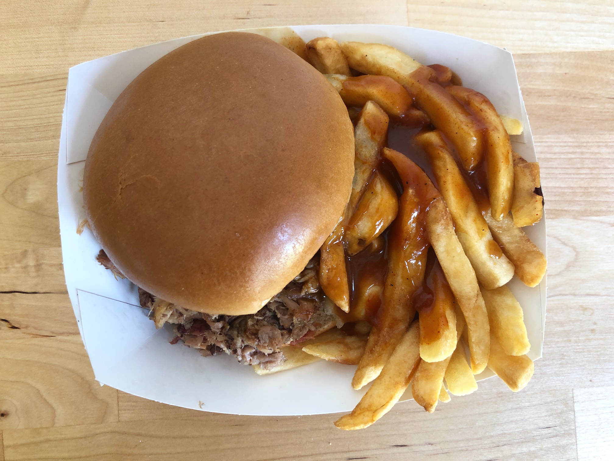 A pulled pork sandwich from Wood N' Hog sits in a white paper tray beside a side of fries doused in barbecue sauce. Photo by Alyssa Buckley.