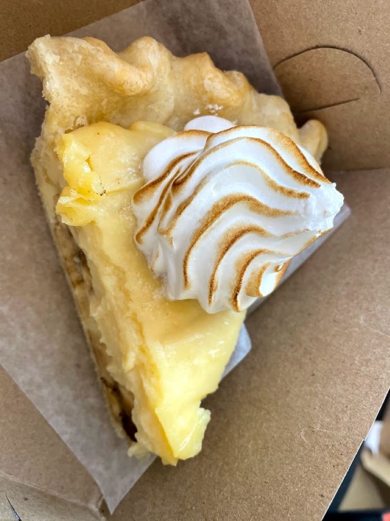 In a brown takeout bow, there is a triangular piece of banana cream pie which is yellow with a white frosting that has little brown streaks from charring. Photo by Remington Rock.