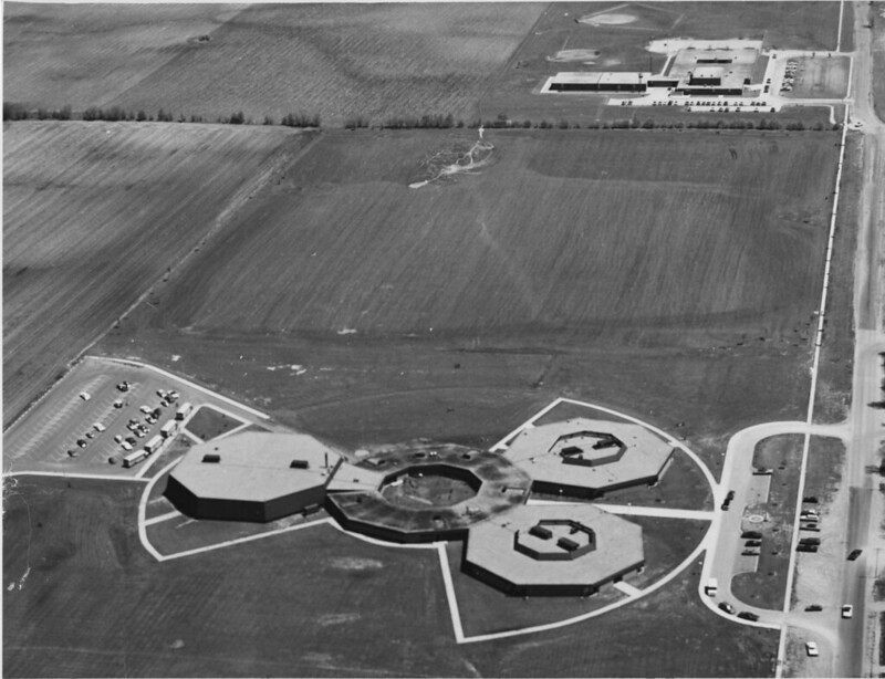 A black and white aerial photo of a building with four hexagonal shaped components. It is in the middle of a large field. Another building is at the top of the photo. Photo from Urbana Free Library Facebook page.
