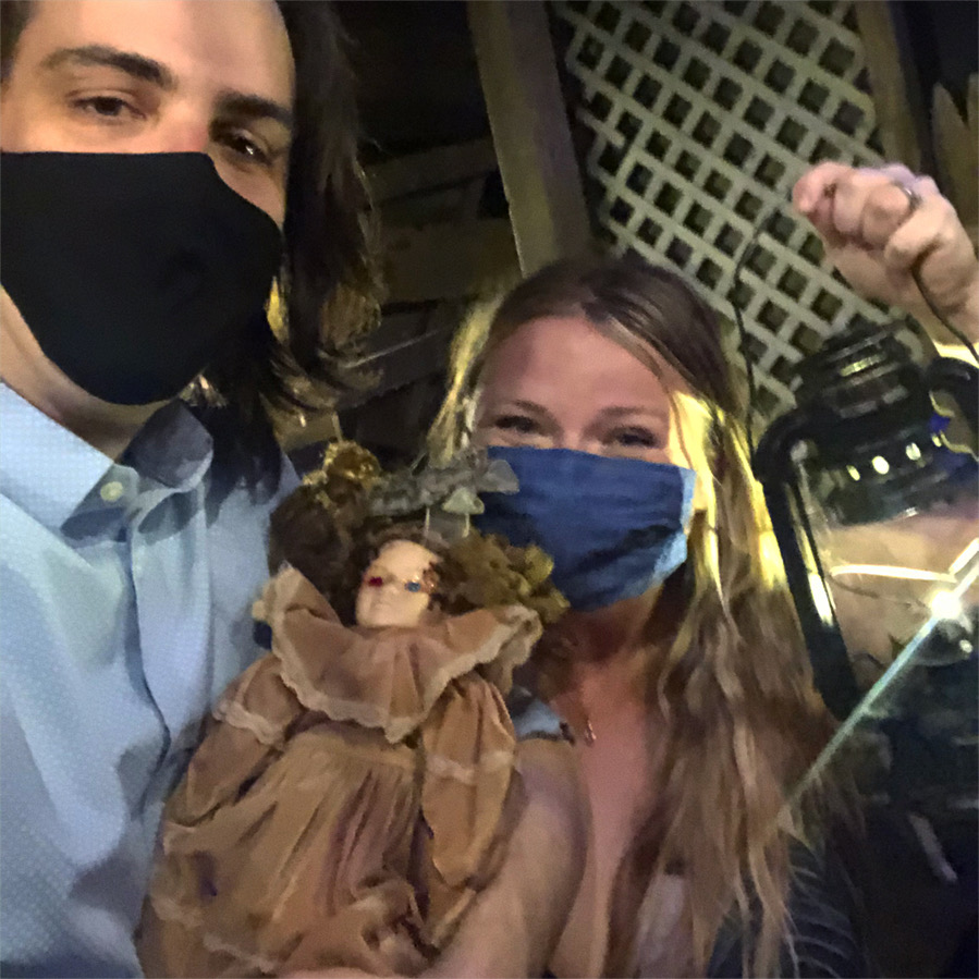 Photo of the author and her husband after solving Revenge of the Cabin. Both participants are masked, and the man is holding a creepy doll, and the woman is holding a lantern. Photo by Alyssa Buckley.