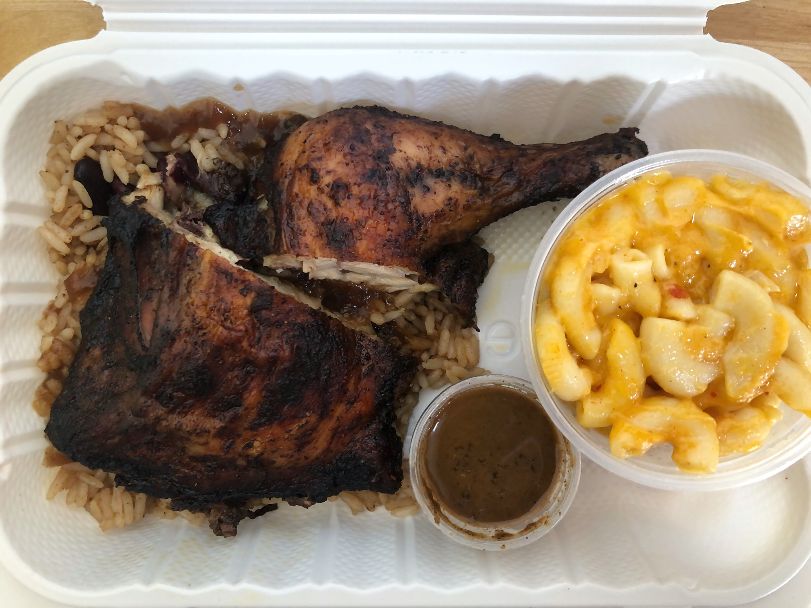 In a white takeout container, there is Caribbean Grill's Flavor Box lunch with two pieces of chicken with a dark skin over light brown rice with a plastic cup of macaroni beside a tiny cup of jerk sauce. Photo by Alyssa Buckley.