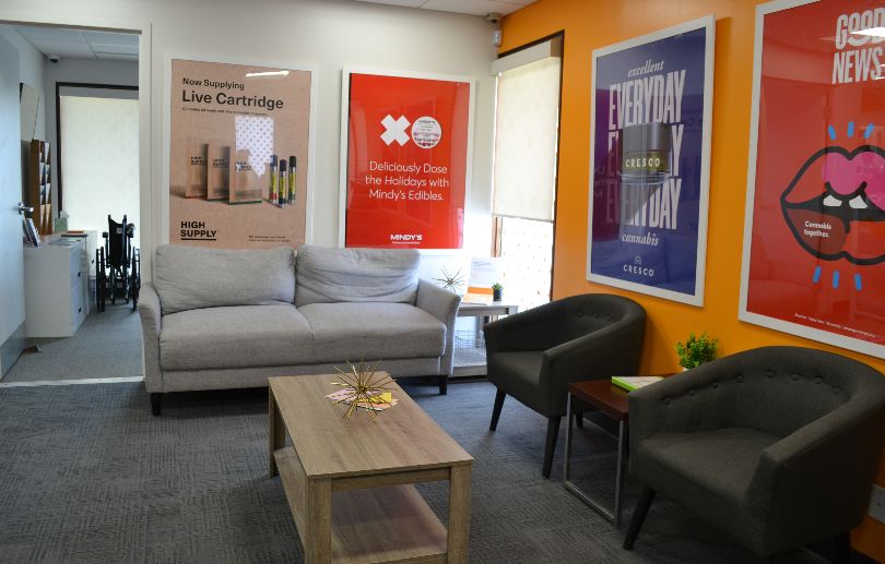 In the lobby of Sunnyside Champaign, there is a beige couch and two gray midcentury modern chairs facing a long, rectangular coffee table. There are cannabis product advertisements framed in poster frames on white walls. Photo by Alyssa Buckley.