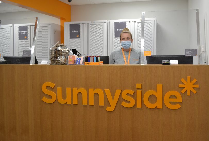 At a brown wooden counter, there is a Sunnyside wellness advisor in a mask with an orange lanyard and a blonde topknot bun ready to serve customers. Photo by Alyssa Buckley.