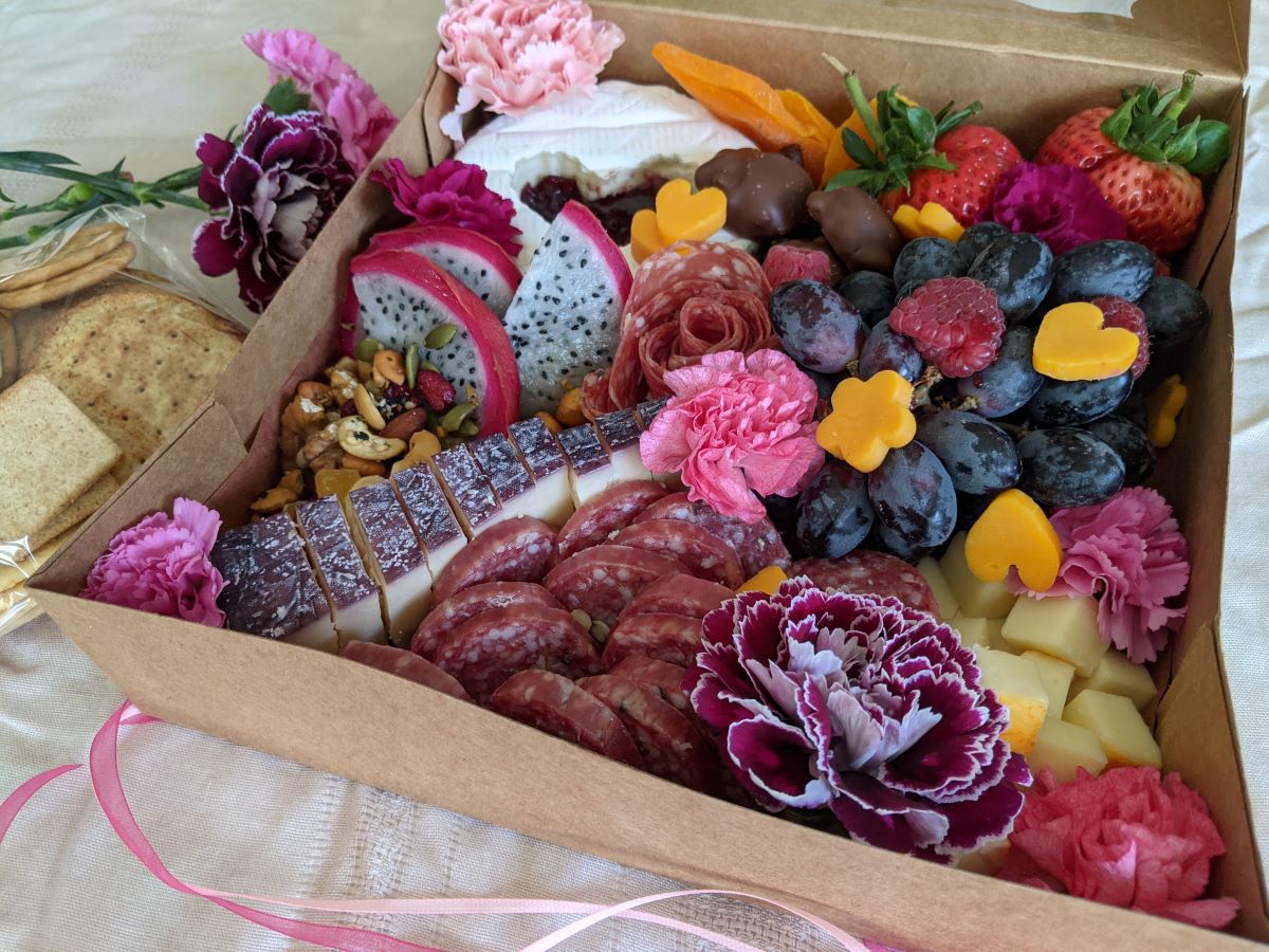 A brown cardboard box with various meats, cheeses, and fruits inside and decorated with carnation flowers.  To the left of the box is a pack of crackers.  Pink ribbons are at the bottom of the photo. Photo by Tias Paul.