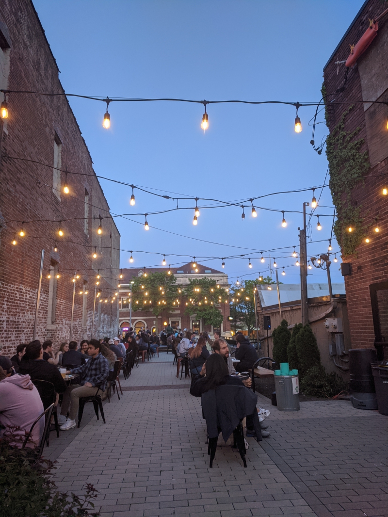 An outdoor patio between two brick buildings. String lights hang between the buildings. There are two rows of tables, each filled with people. Photo by Tom Ackerman.