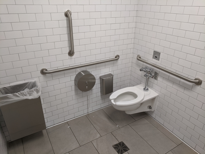A white porcelain toilet is surrounded by white tiled walls. There are metal handles on the wall, as well as a waste dispenser and toilet paper holder. Photo by Tom Ackerman.
